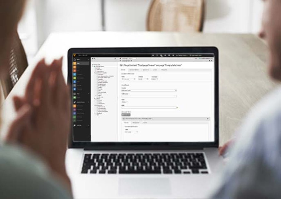 Es sind zwei Personen von hinten (verschwommen) zu sehen, die auf einen Laptop schauen, auf dem das Backend einer TYPO3 Website zu sehen ist.